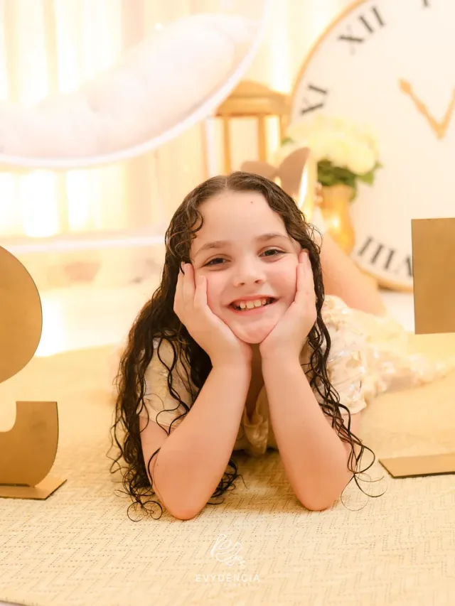 Menina sorridente deitada no tapete redondo dourado entre os números do Ano Novo com o grande relógio ao fundo