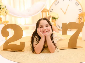 Menina sorridente deitada no tapete redondo dourado entre os números do Ano Novo com o grande relógio ao fundo