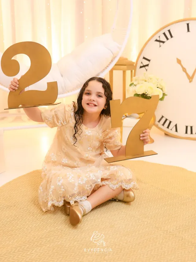 Cenário de Ano Novo no Estúdio Evydência: menina em vestido champanhe com números dourados e grande relógio às 11:55