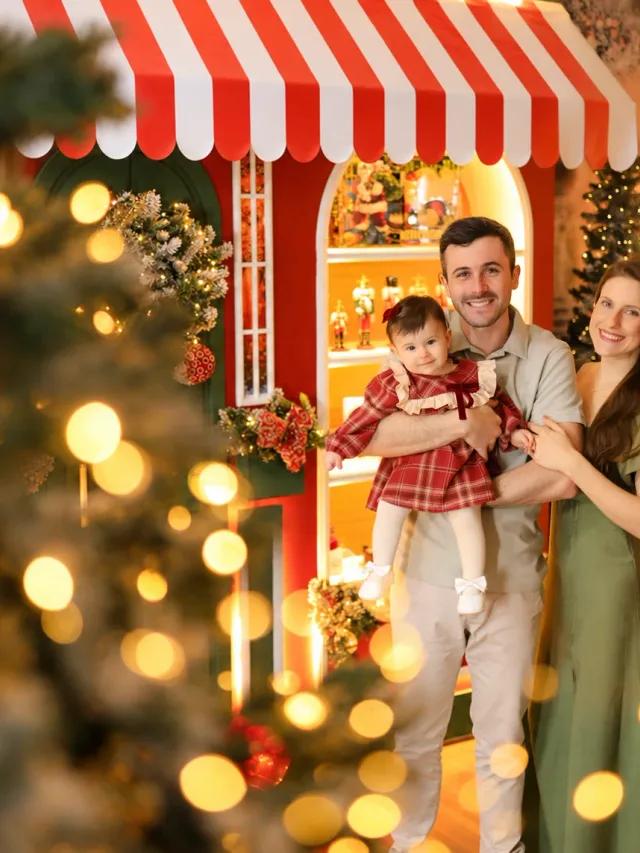 Ensaio de família no Natal: pai, mãe e bebê de vermelho no toldo da loja de brinquedos da Vila de Natal em Tijucas