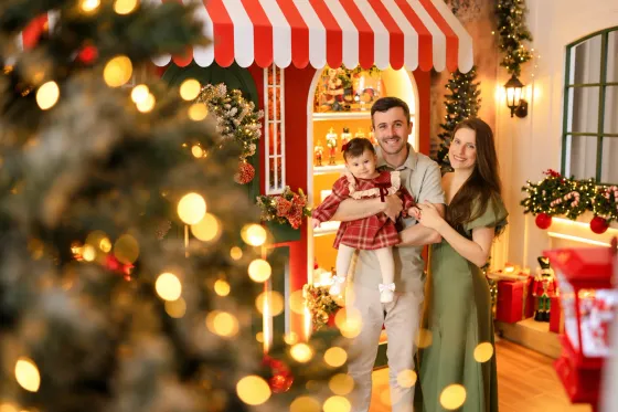 Ensaio de família no Natal: pai, mãe e bebê de vermelho no toldo da loja de brinquedos da Vila de Natal em Tijucas