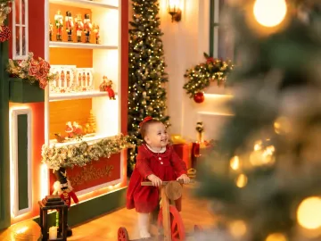 Bebê em vestido xadrez andando no triciclo de madeira vintage em frente à vitrine iluminada da Vila de Natal