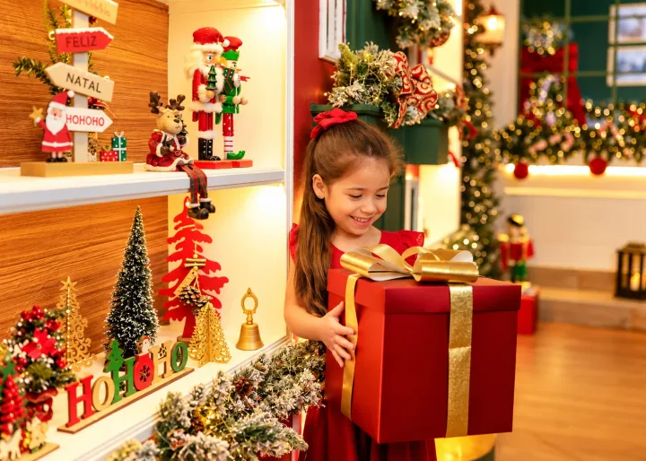 Menina de vermelho segurando grande presente natalino com laço dourado na prateleira da Vila de Natal