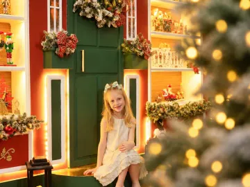 Menina em vestido marfim sentada no degrau do portal verde com guirlanda na Vila de Natal em Tijucas