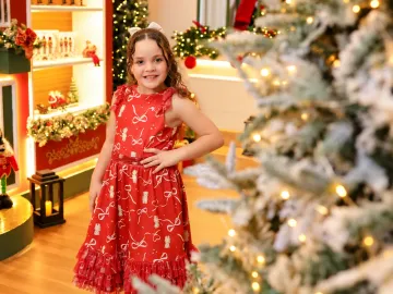 Menina em vestido vermelho de festa sorrindo em frente à Vila de Natal e pinheiro iluminado em Tijucas
