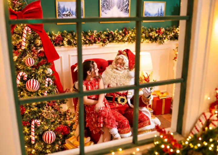 Papai Noel com figurino clássico e barba real conversando com menina na poltrona vermelha visto pela janela