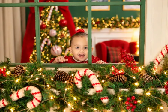 Bebê sorridente espiando pela janela verde decorada com guirlanda, pinhas e bengalas doces no Estúdio Evydência