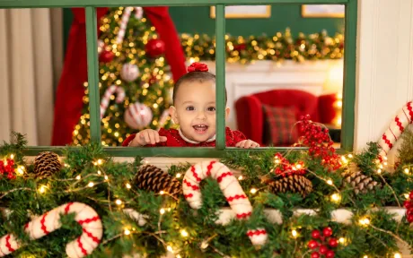 Bebê sorridente espiando pela janela verde decorada com guirlanda, pinhas e bengalas doces no Estúdio Evydência