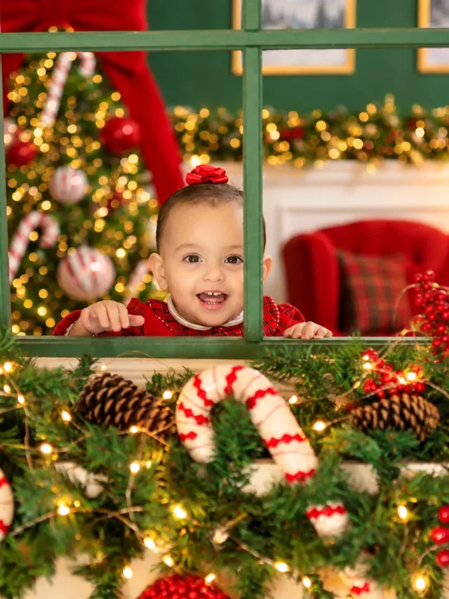 Bebê sorridente espiando pela janela verde decorada com guirlanda, pinhas e bengalas doces no Estúdio Evydência