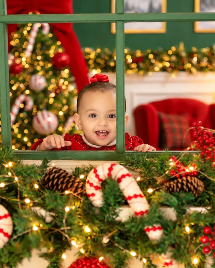Bebê sorridente espiando pela janela verde decorada com guirlanda, pinhas e bengalas doces no Estúdio Evydência