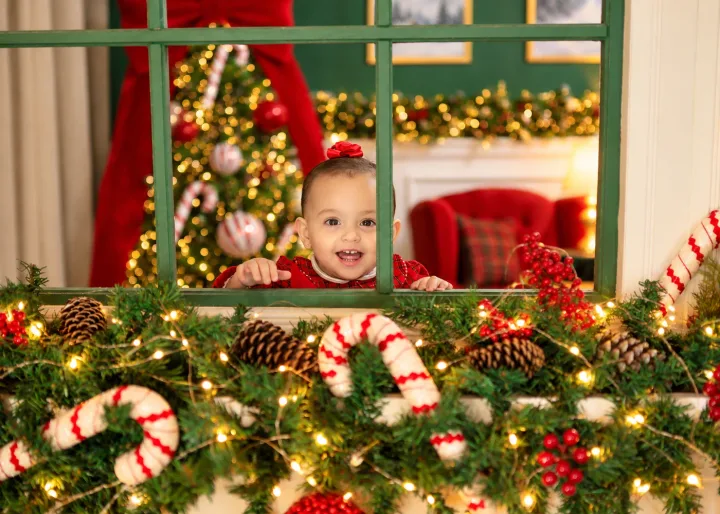 Bebê sorridente espiando pela janela verde decorada com guirlanda, pinhas e bengalas doces no Estúdio Evydência