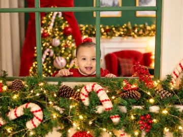 Bebê sorridente espiando pela janela verde decorada com guirlanda, pinhas e bengalas doces no Estúdio Evydência
