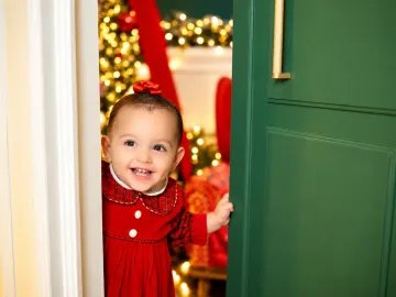 Bebê espiando pela porta verde da Vila de Natal com sorriso alegre e olhar curioso no Estúdio Evydência